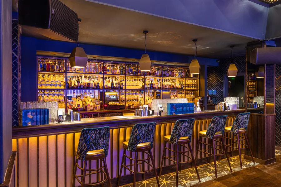 Bar z podświetloną półką pełną butelek, granatową boazerią i rzędem stołków